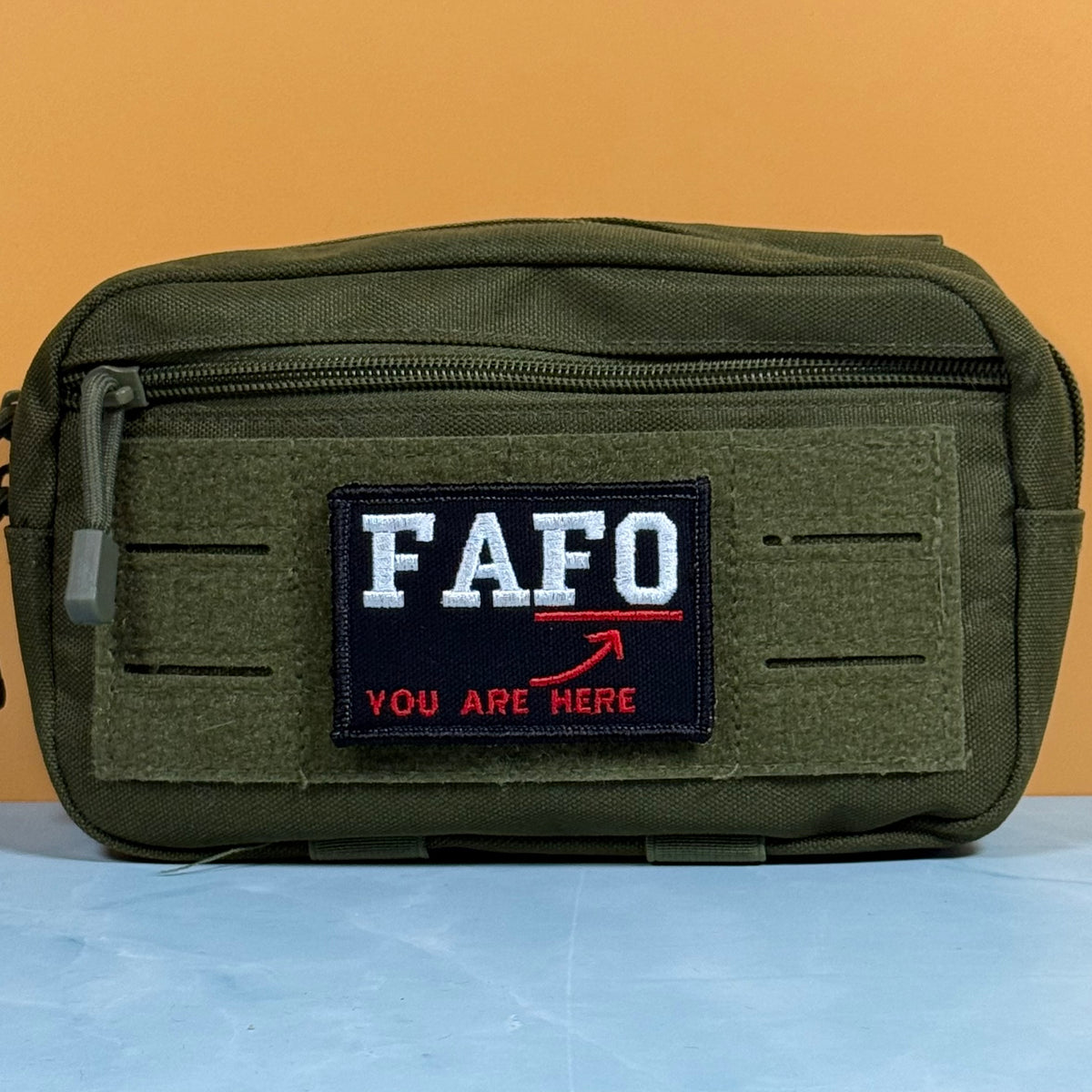 An olive green tactical pouch with a front zipper and a custom embroidered patch reading 'FAFO YOU ARE HERE' in white and red, set against a solid orange background.