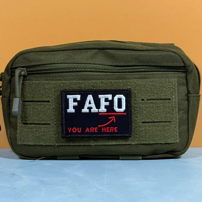 An olive green tactical pouch with a front zipper and a custom embroidered patch reading 'FAFO YOU ARE HERE' in white and red, set against a solid orange background.