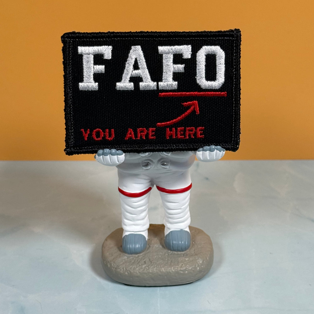 A novelty astronaut figurine holding a black rectangular patch that reads 'FAFO YOU ARE HERE' in white and red embroidery, set against a solid orange background and light gray surface.