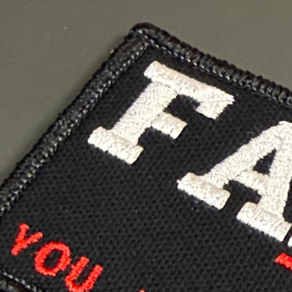 Close-up of a black embroidered patch with white and red lettering on a dark gray background.