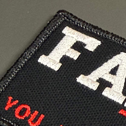 Close-up of a black embroidered patch with white and red lettering on a dark gray background.