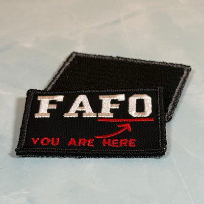 A black embroidered patch with 'FAFO' in white letters and 'YOU ARE HERE' in red, plus a red arrow, shown on a light gray surface with a second black patch behind it.