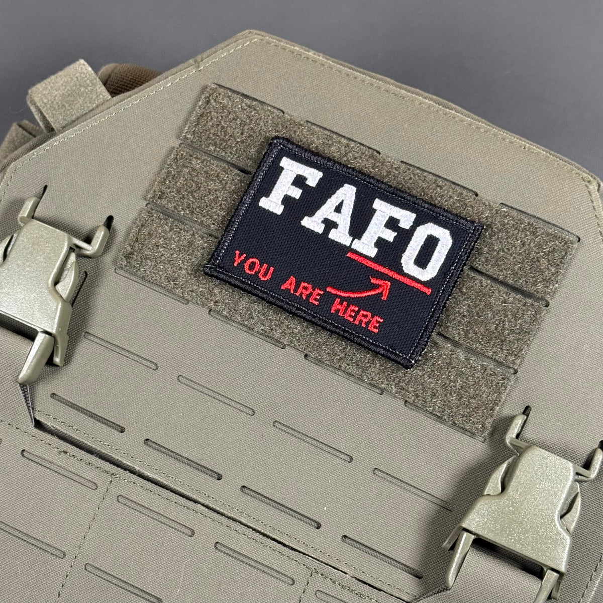 A tactical patch with the text 'FAFO YOU ARE HERE' in white and red on a black background, attached to a tan MOLLE webbing panel on a matching tan tactical bag.