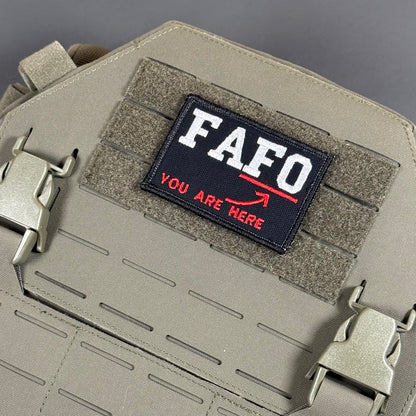 A tactical patch with the text 'FAFO YOU ARE HERE' in white and red on a black background, attached to a tan MOLLE webbing panel on a matching tan tactical bag.