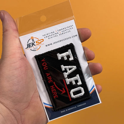 A hand holding a clear plastic blister pack containing an embroidered patch. The patch is black with large white 'FAFO' lettering and red text reading 'YOU ARE HERE' with a red arrow.