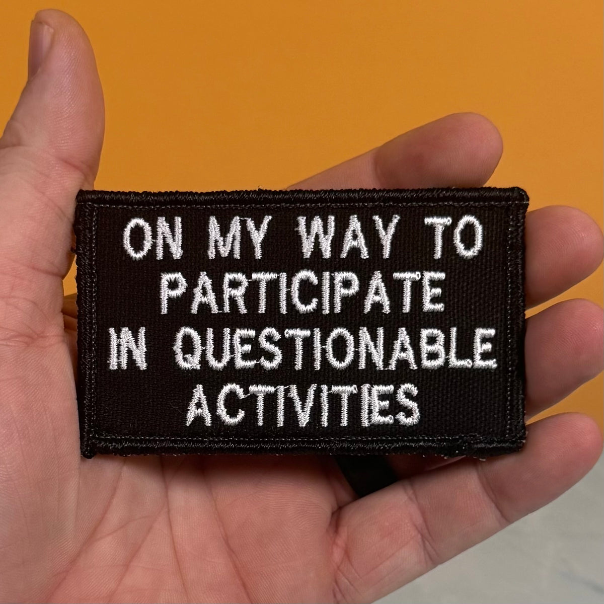 A hand holding a rectangular black embroidered patch with white text that reads, 'ON MY WAY TO PARTICIPATE IN QUESTIONABLE ACTIVITIES' against a solid orange background.