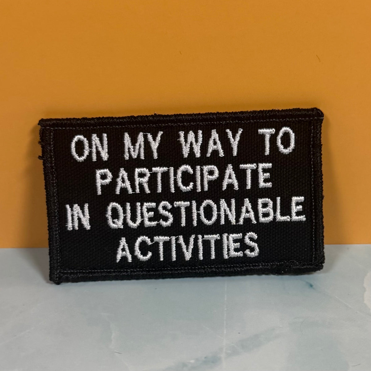 A rectangular black embroidered patch with white text reading 'ON MY WAY TO PARTICIPATE IN QUESTIONABLE ACTIVITIES' against a mustard yellow background and light gray surface.