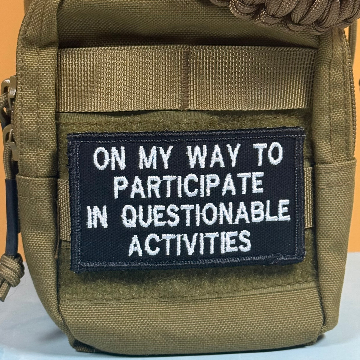 An olive drab tactical pouch with a black embroidered patch that reads 'ON MY WAY TO PARTICIPATE IN QUESTIONABLE ACTIVITIES' in white text.