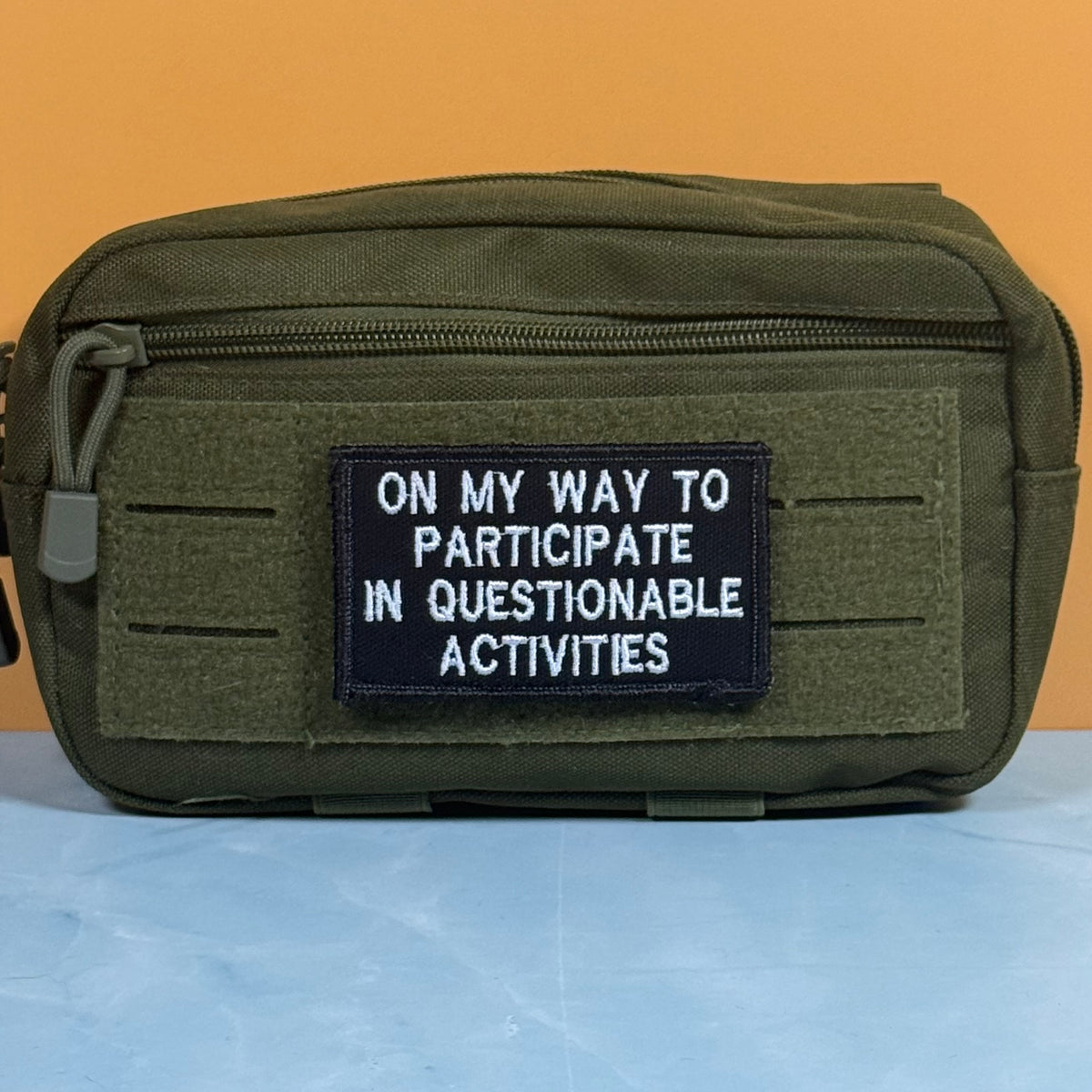 An olive green tactical pouch with a front zipper and a black rectangular patch that reads 'ON MY WAY TO PARTICIPATE IN QUESTIONABLE ACTIVITIES' in white text, set against a solid orange background.