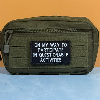 An olive green tactical pouch with a front zipper and a black rectangular patch that reads 'ON MY WAY TO PARTICIPATE IN QUESTIONABLE ACTIVITIES' in white text, set against a solid orange background.