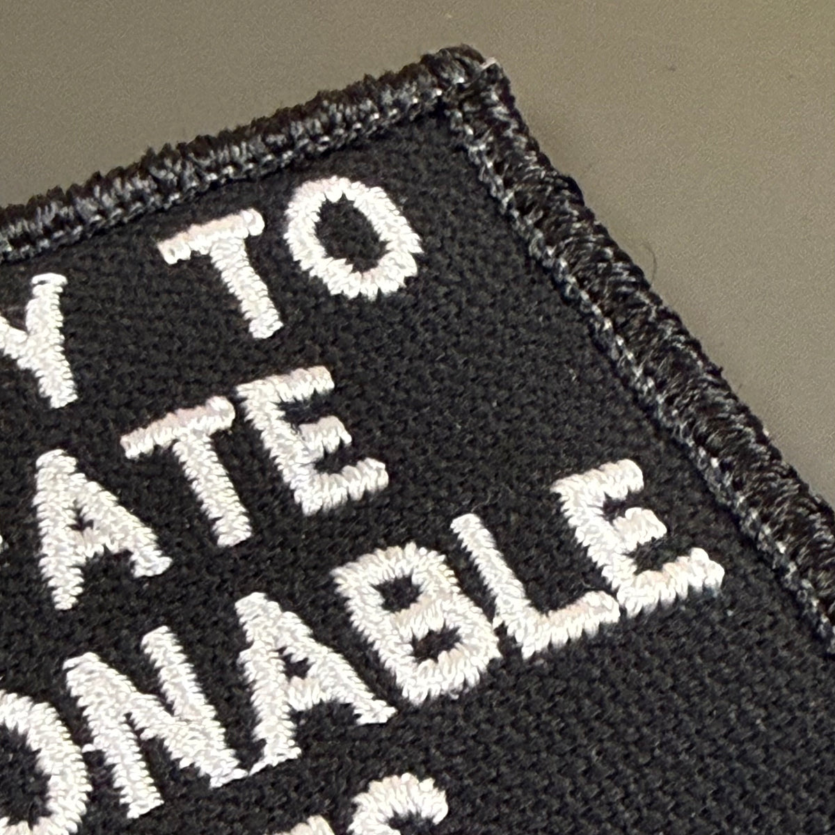 Close-up of a black embroidered patch with white text reading 'WAY TO ATE NONABLE' on a plain taupe background.