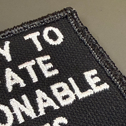 Close-up of a black embroidered patch with white text reading 'WAY TO ATE NONABLE' on a plain taupe background.