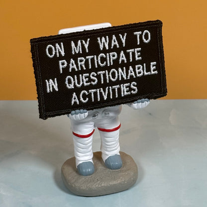 An embroidered patch with the text 'ON MY WAY TO PARTICIPATE IN QUESTIONABLE ACTIVITIES' in white on a dark brown background, held by a small astronaut figurine on a gray base against an orange and gray background.