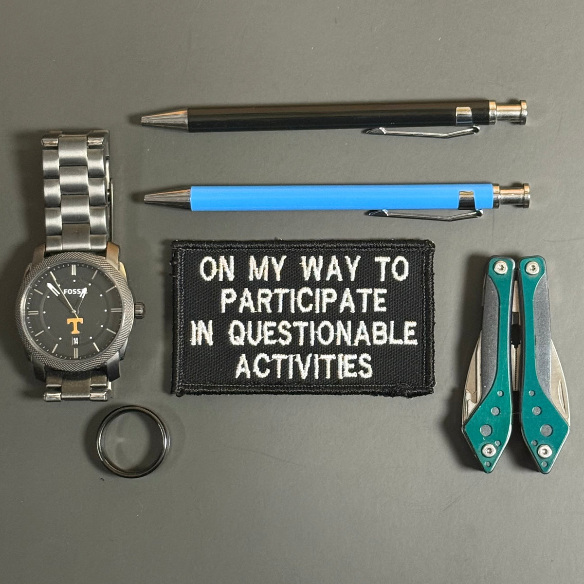 A flat-lay arrangement of everyday carry accessories on a gray background, including a stainless steel wrist watch, a black pen, a blue pen, a black embroidered patch with white text reading 'ON MY WAY TO PARTICIPATE IN QUESTIONABLE ACTIVITIES', a folding pocket knife with teal handles, and a black ring.