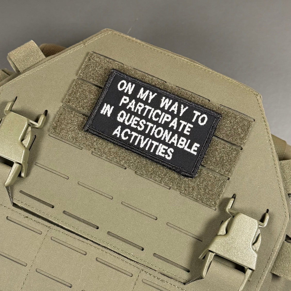 A black rectangular embroidered patch with white text reading 'ON MY WAY TO PARTICIPATE IN QUESTIONABLE ACTIVITIES' attached to a tan tactical plate carrier vest.