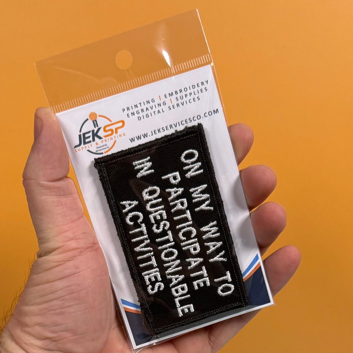 A hand holds a packaged black embroidered patch with white text reading 'ON MY WAY TO PARTICIPATE IN QUESTIONS ONLY.' The backing card shows the JEKSP embroidery supplies logo and website, against a solid orange background.