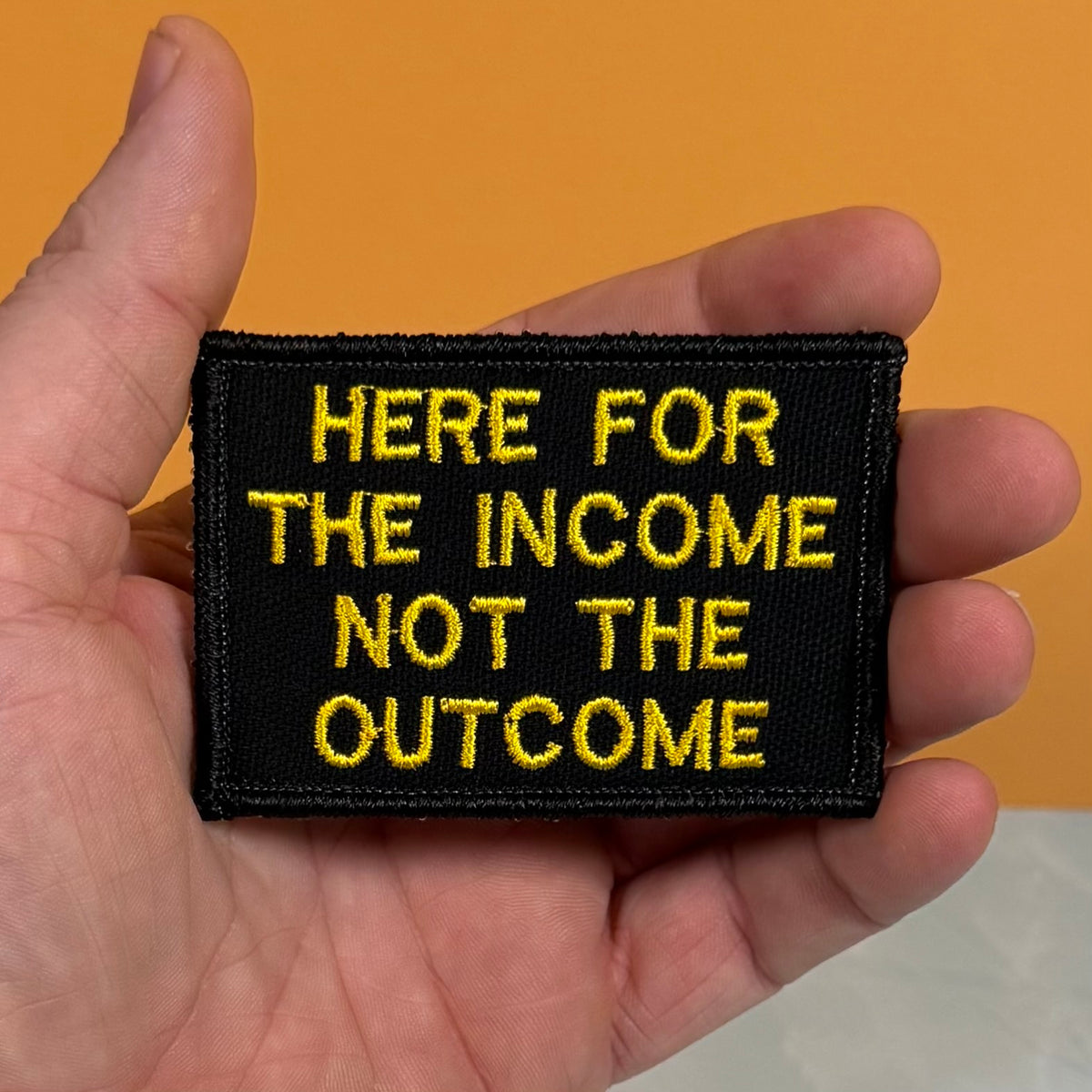 A hand holding a rectangular black embroidered patch with yellow text that reads 'HERE FOR THE INCOME NOT THE OUTCOME' against an orange background.