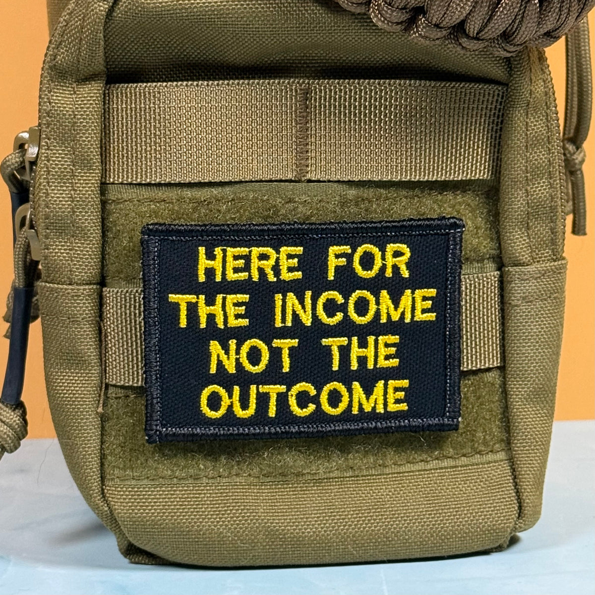 An olive green tactical pouch with a black rectangular patch featuring yellow embroidered text that reads 'HERE FOR THE INCOME NOT THE OUTCOME'.