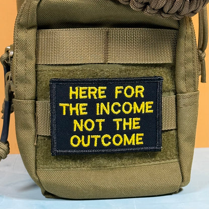 An olive green tactical pouch with a black rectangular patch featuring yellow embroidered text that reads 'HERE FOR THE INCOME NOT THE OUTCOME'.