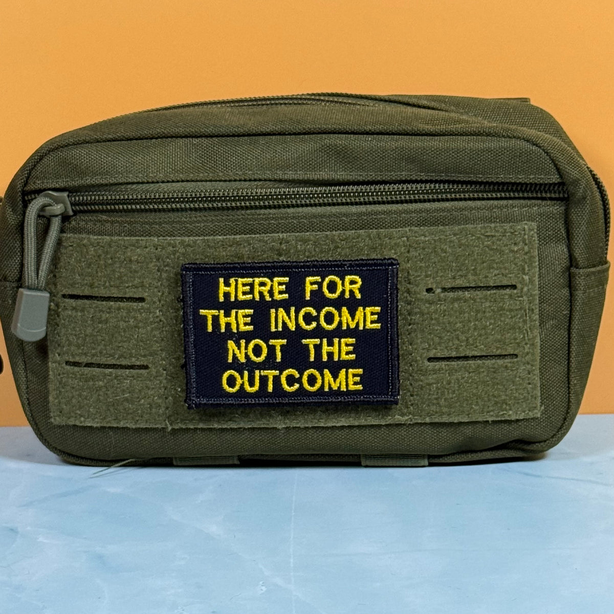 An olive green tactical pouch with a black rectangular patch featuring yellow embroidered text that reads 'HERE FOR THE INCOME NOT THE OUTCOME'. The pouch is displayed on a light blue surface against a solid orange background.