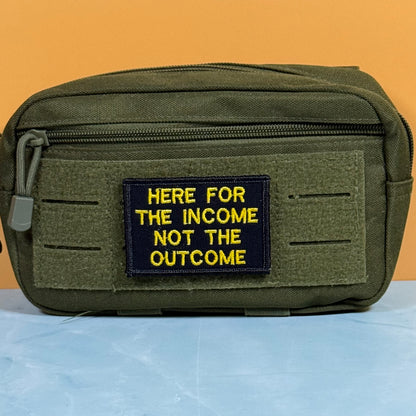 An olive green tactical pouch with a black rectangular patch featuring yellow embroidered text that reads 'HERE FOR THE INCOME NOT THE OUTCOME'. The pouch is displayed on a light blue surface against a solid orange background.