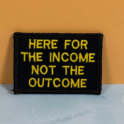 A rectangular black embroidered patch with yellow text reading 'HERE FOR THE INCOME NOT THE OUTCOME', photographed against an orange wall and light gray surface.