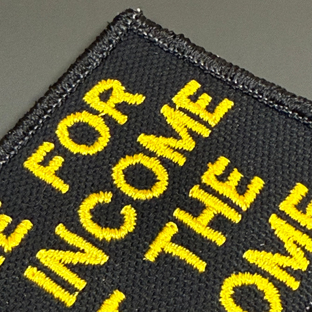 Close-up of a black embroidered patch with bold yellow text reading 'FOR INCOME TO THE HOME' on a dark gray background.