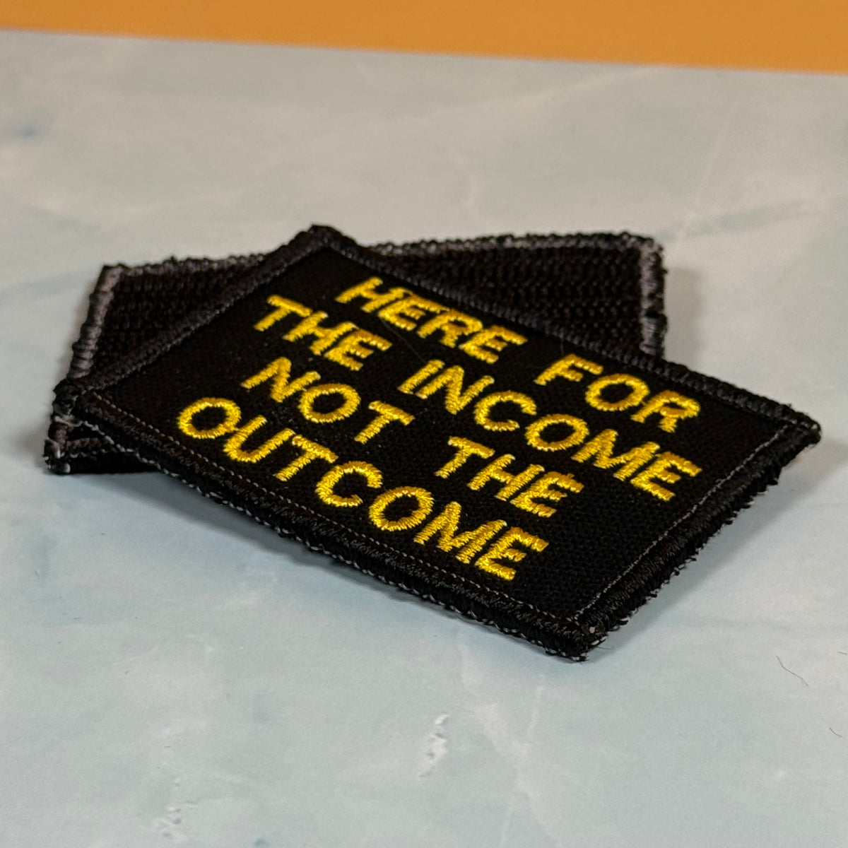 Two black embroidered patches with yellow text reading 'HERE FOR THE INCOME NOT THE OUTCOME' stacked on a light gray surface.