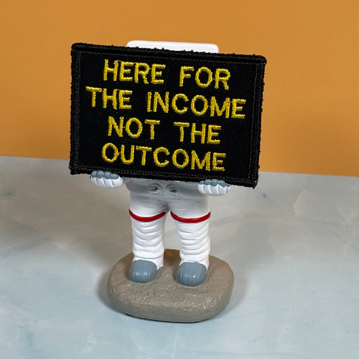 An embroidered patch with the text 'HERE FOR THE INCOME NOT THE OUTCOME' in yellow on a black background, held by a small astronaut figurine on a gray base against an orange and light gray background.