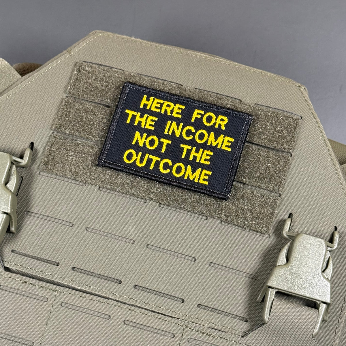 A rectangular black tactical patch with yellow embroidered text reading 'HERE FOR THE INCOME NOT THE OUTCOME', attached to a tan webbing plate on a matching tan tactical vest or backpack.