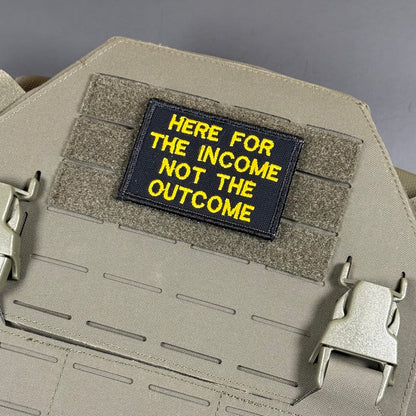 A rectangular black tactical patch with yellow embroidered text reading 'HERE FOR THE INCOME NOT THE OUTCOME', attached to a tan webbing plate on a matching tan tactical vest or backpack.