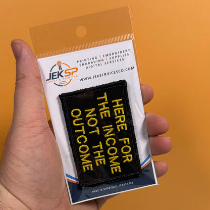 A hand holds a packaged black embroidered patch with yellow text reading 'HERE FOR THE INCOME NOT THE OUTCOME' against an orange background. The card backing shows the JEKSP brand and website.