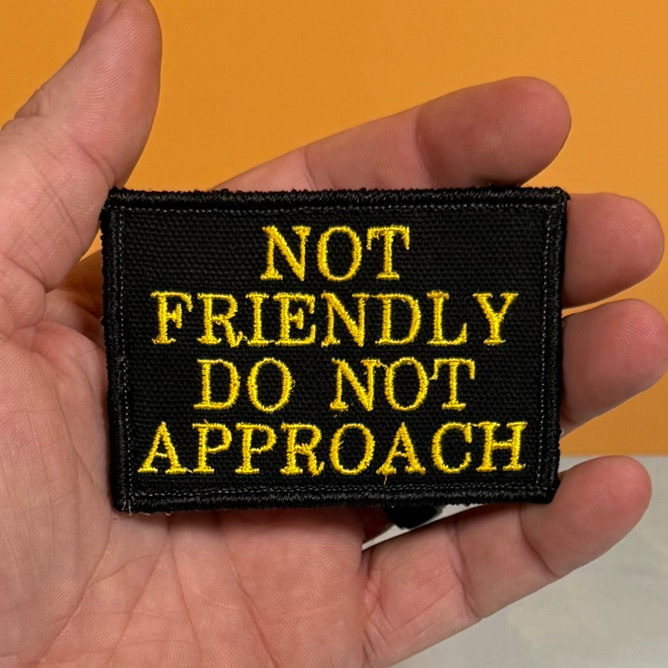 A rectangular black embroidered patch with yellow text reading 'NOT FRIENDLY DO NOT APPROACH' held in a person's hand against a solid orange background.