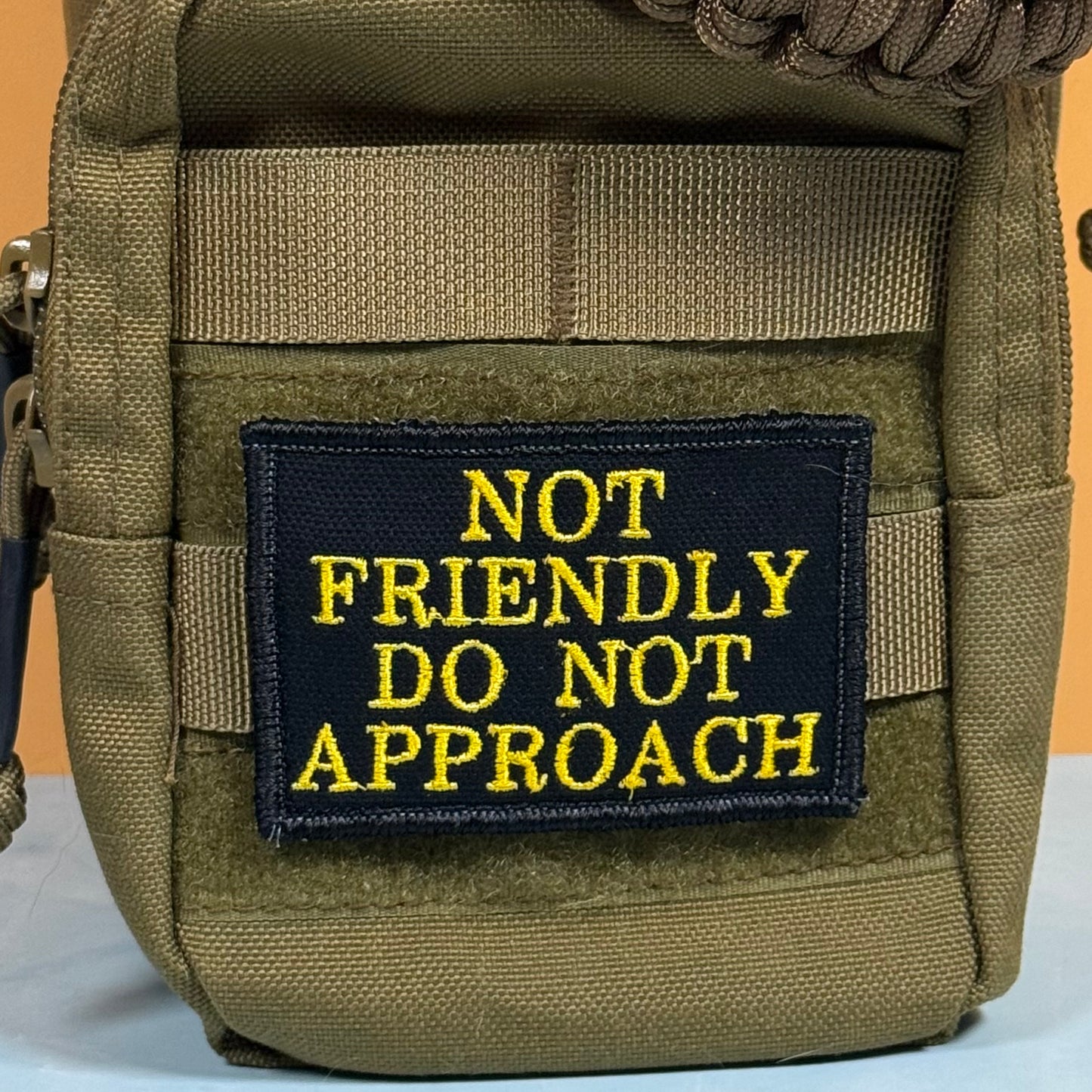 An olive drab tactical pouch featuring a black rectangular patch with yellow embroidered text that reads 'NOT FRIENDLY DO NOT APPROACH'.