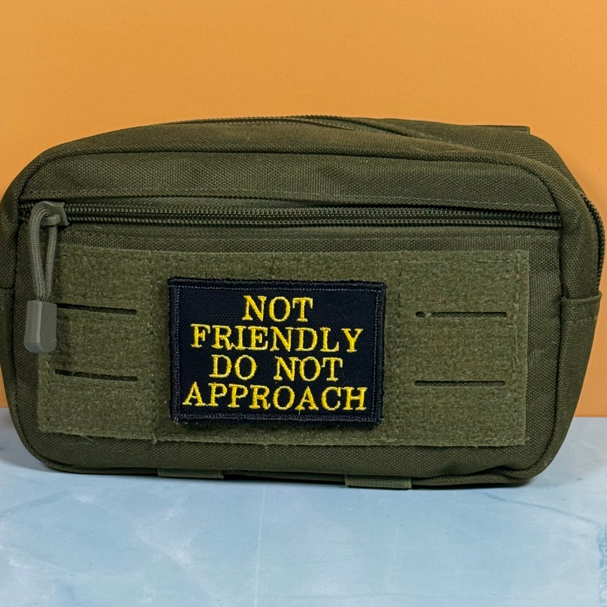 An olive green tactical pouch with a black rectangular patch featuring yellow embroidered text that reads 'NOT FRIENDLY DO NOT APPROACH'. The pouch is shown on a light blue surface against a solid orange background.