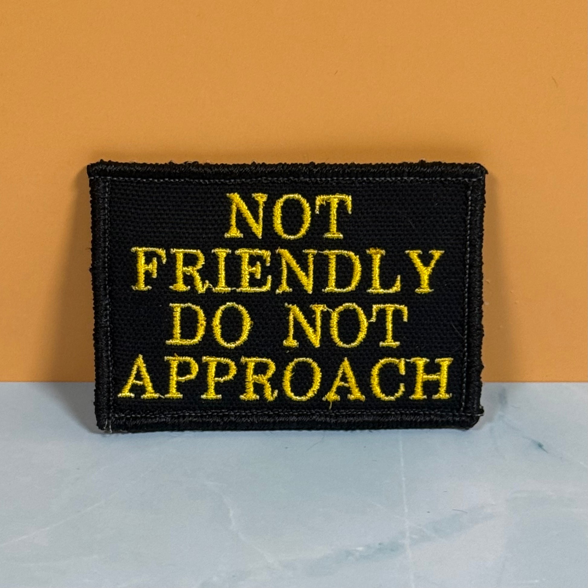 A rectangular black embroidered patch with yellow text reading 'NOT FRIENDLY DO NOT APPROACH', photographed standing upright against an orange wall on a light marble-like surface.