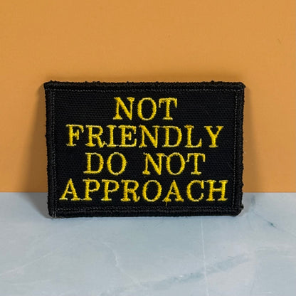 A rectangular black embroidered patch with yellow text reading 'NOT FRIENDLY DO NOT APPROACH', photographed standing upright against an orange wall on a light marble-like surface.