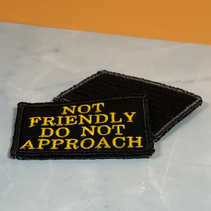 Two black embroidered patches with yellow text reading 'NOT FRIENDLY DO NOT APPROACH' on a light gray surface with an orange background.