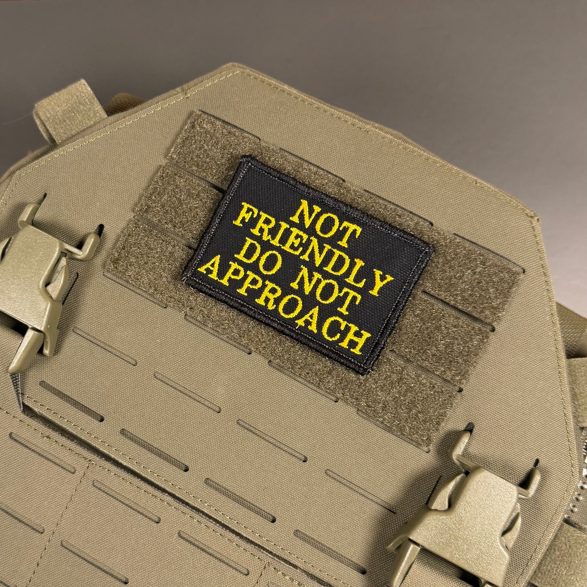 A tactical patch with the text 'NOT FRIENDLY DO NOT APPROACH' in yellow embroidery on a black background, attached to a tan MOLLE plate carrier vest.
