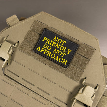 A tactical patch with the text 'NOT FRIENDLY DO NOT APPROACH' in yellow embroidery on a black background, attached to a tan MOLLE plate carrier vest.