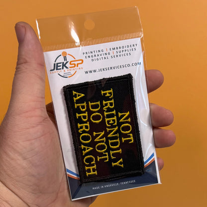 A hand holds a packaged embroidered patch that reads 'NOT FRIENDLY DO NOT APPROACH' in yellow on a black background. The package header shows the Jeksp brand and text for printing, embroidery, supplies, and digital services, with an orange background behind the hand.