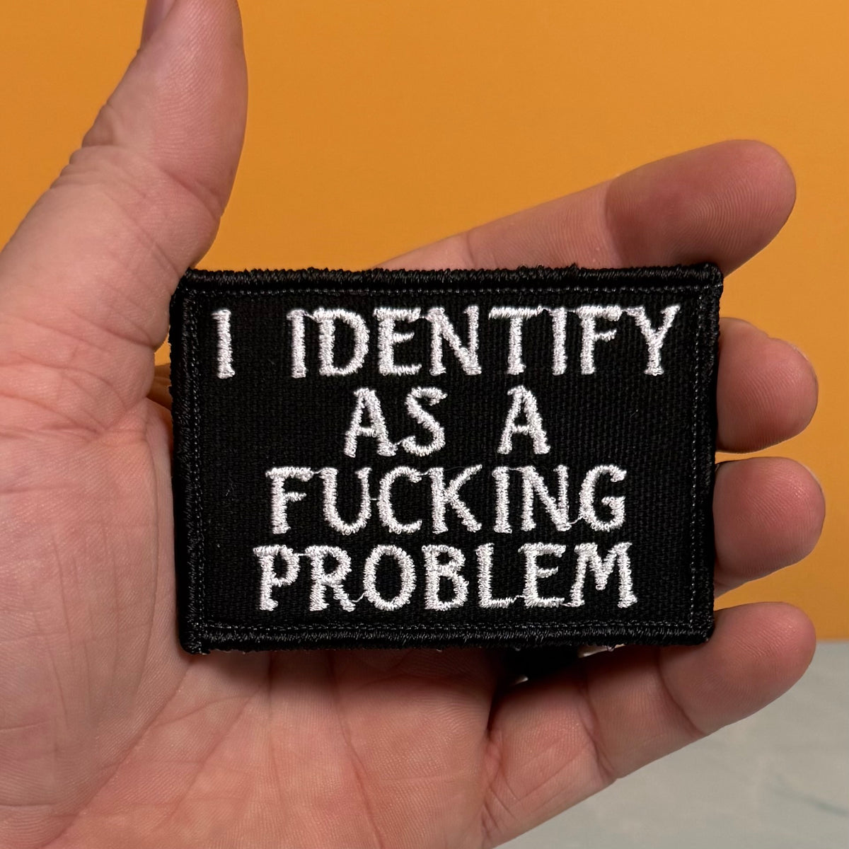 A rectangular black embroidered patch with white text reading 'I IDENTIFY AS A FUCKING PROBLEM' held in a person's hand against a solid orange background.