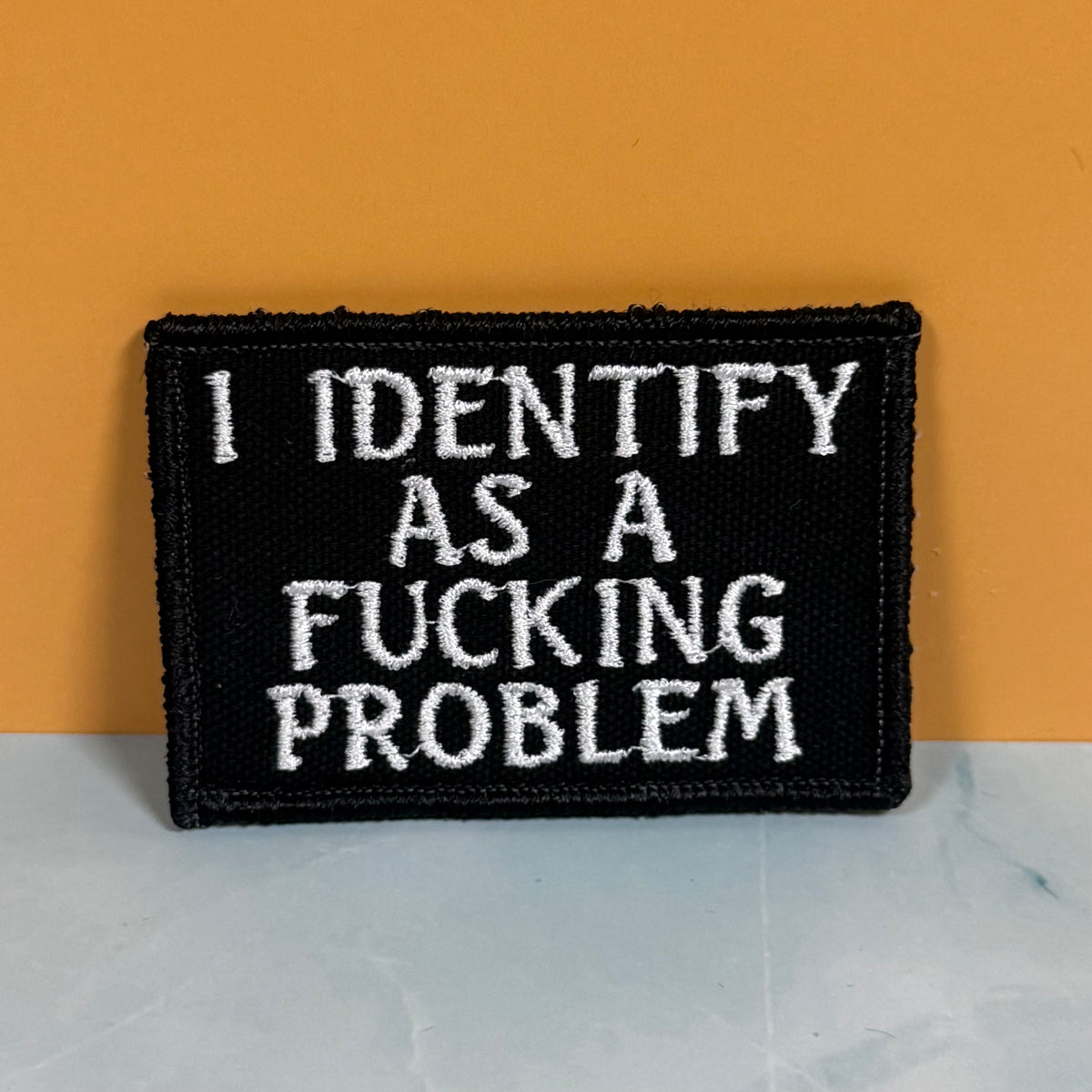 A rectangular black embroidered patch with white text reading 'I IDENTIFY AS A FUCKING PROBLEM,' photographed against a mustard-yellow background and a pale marble-like surface.
