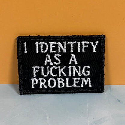 A rectangular black embroidered patch with white text reading 'I IDENTIFY AS A FUCKING PROBLEM,' photographed against a mustard-yellow background and a pale marble-like surface.