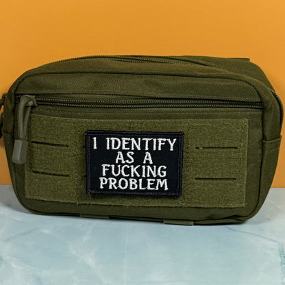 An olive green tactical pouch with a black rectangular patch that reads 'I IDENTIFY AS A FUCKING PROBLEM' in white text, set against a solid orange background.