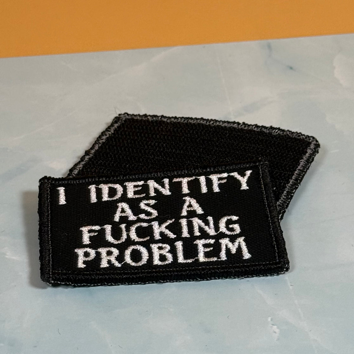 Two black embroidered patches with white text, one reading 'I IDENTIFY AS A FUCKING PROBLEM' and the other partially visible behind it, placed on a light gray marble-like surface with an orange background.