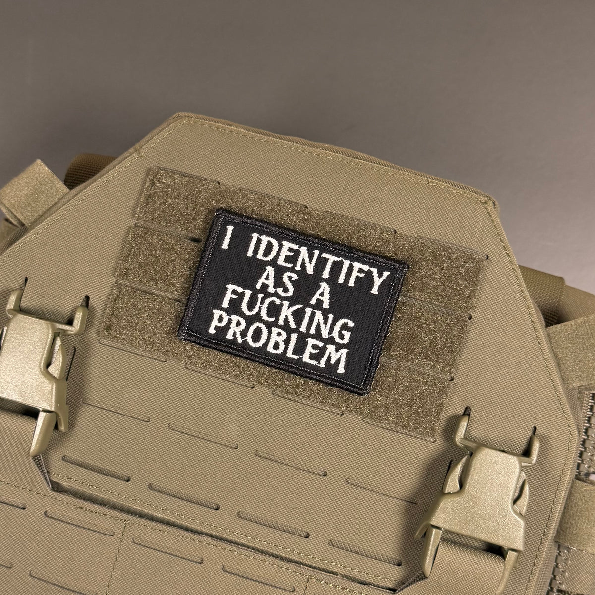 A close-up of a tactical vest or plate carrier in coyote tan, featuring a black rectangular patch with white embroidered text that reads 'I IDENTIFY AS A FUCKING PROBLEM'.