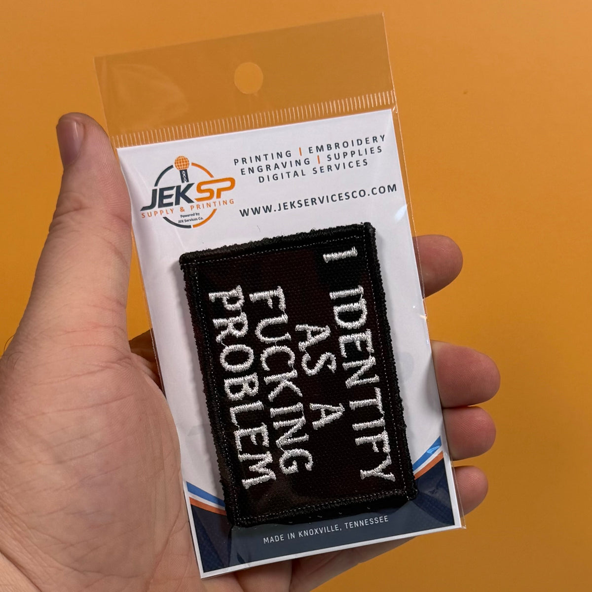 A hand holds a packaged embroidered patch against a solid orange background. The patch reads 'I IDENTIFY AS A FUCKING PROBLEM' in white lettering on a black background, and it is mounted on a white backing card with the JEKSP brand logo and website printed at the top.