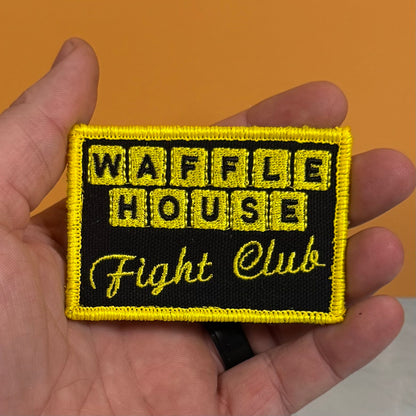 A rectangular embroidered patch with a black background and bright yellow border, held in a hand against an orange background. The patch reads 'WAFFLE HOUSE Fight Club' in yellow embroidery.