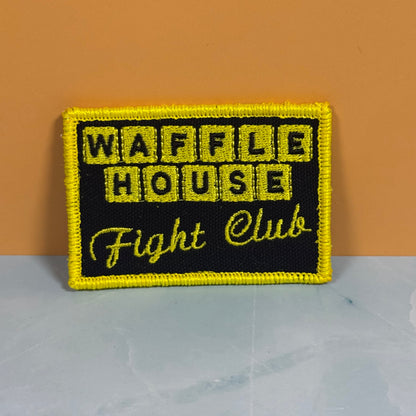 A rectangular embroidered patch with a black background and bright yellow border, reading 'WAFFLE HOUSE Fight Club' in yellow lettering, photographed against an orange wall and pale gray surface.